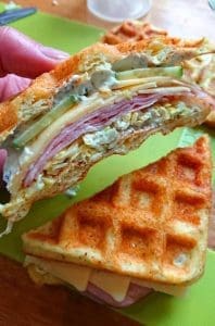 En delad chaffle med skinka, ost och grönsaker i en hand.